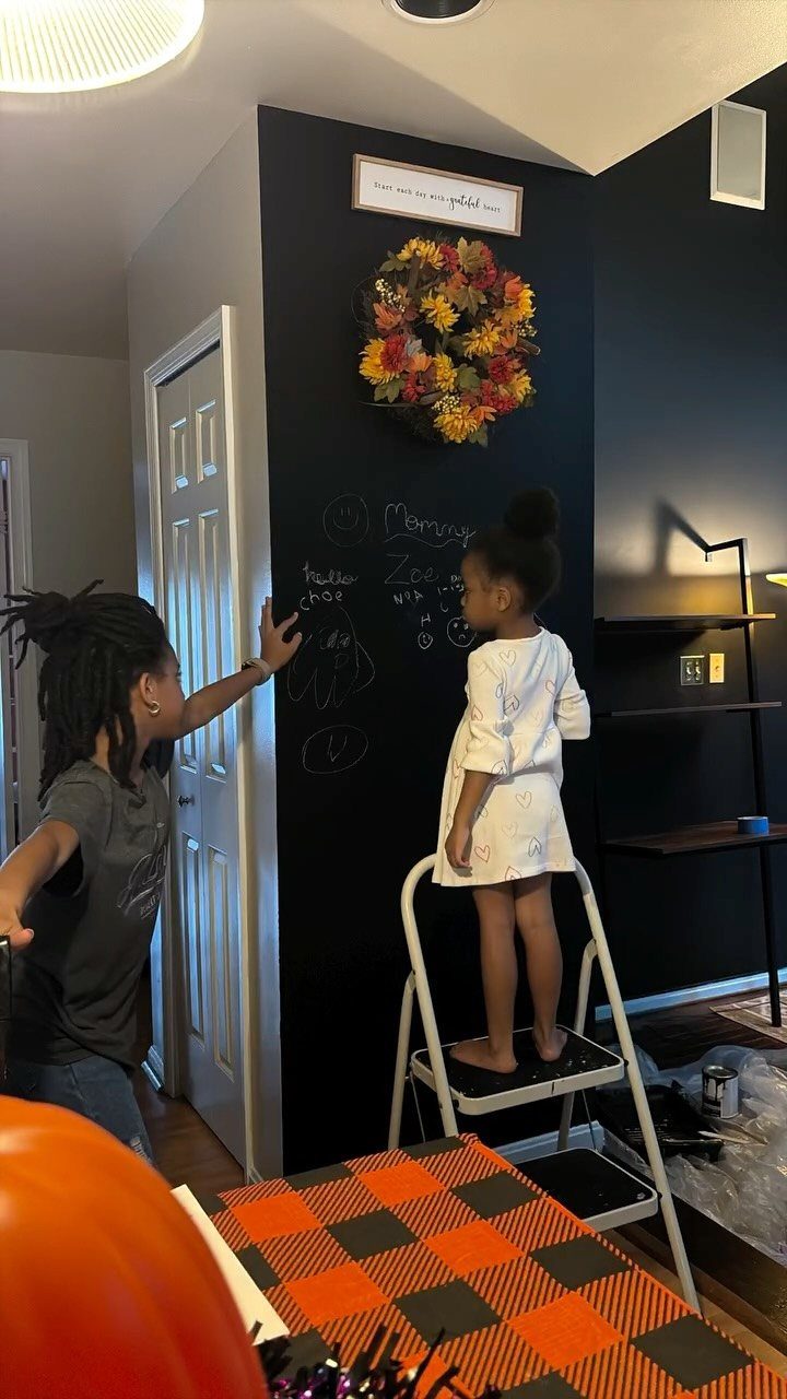 Girl on a step ladder drawing with chalk on a black chalkboard wall