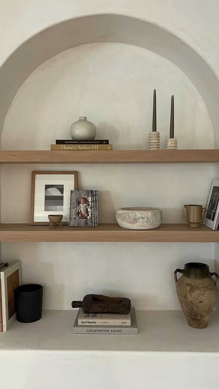Wooden shelves in a white arched niche with neutral decorative pottery and books.