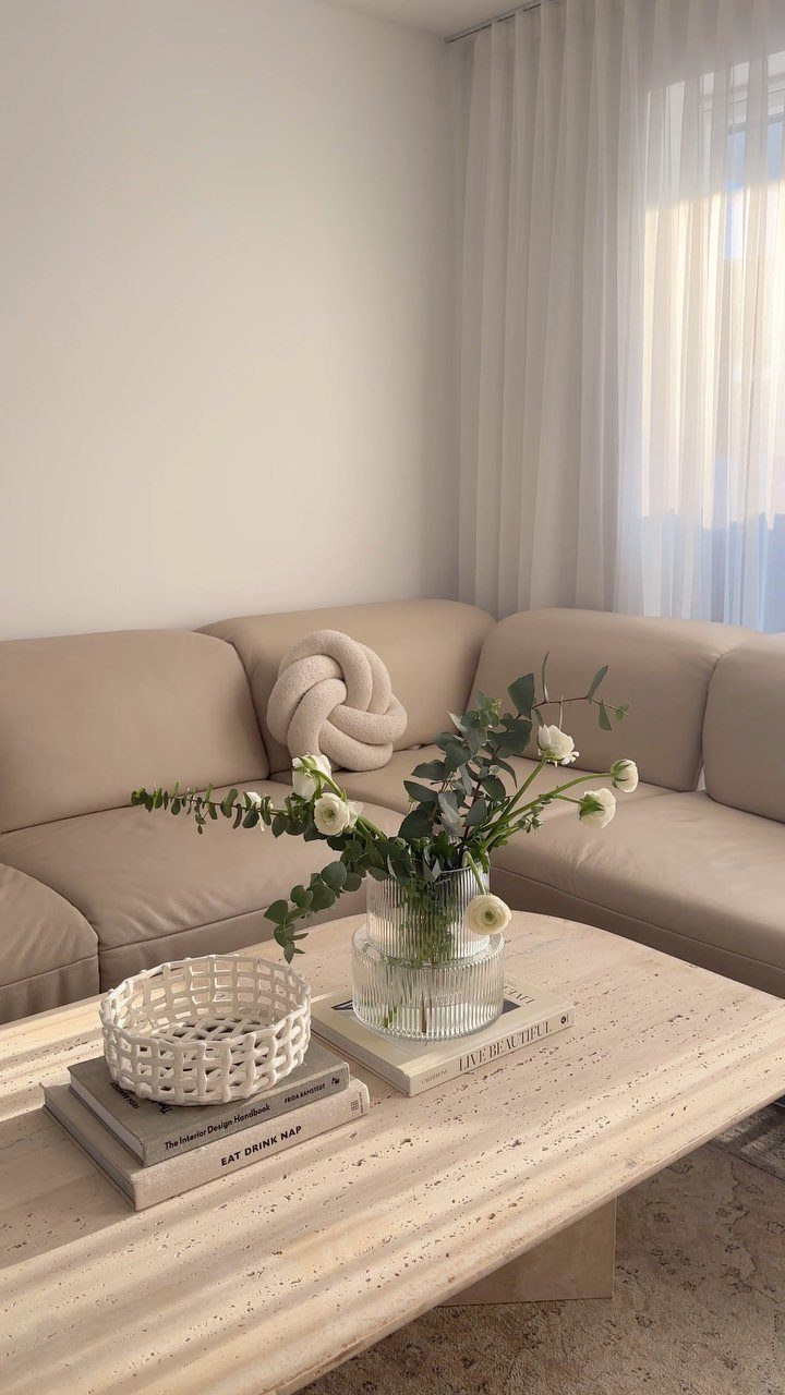 Beige sectional couch behind a travertine coffee table with flowers and books.