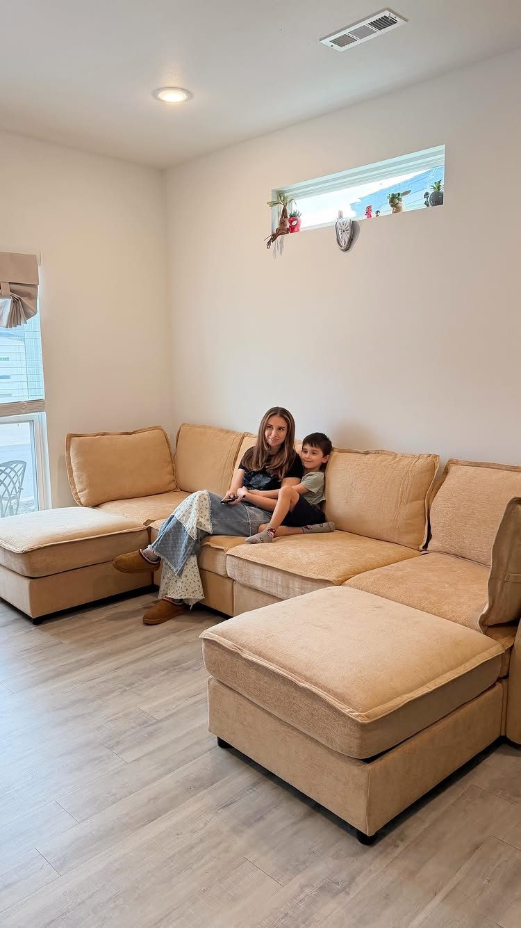 Woman and child sitting on a large beige sectional sofa in a bright room.