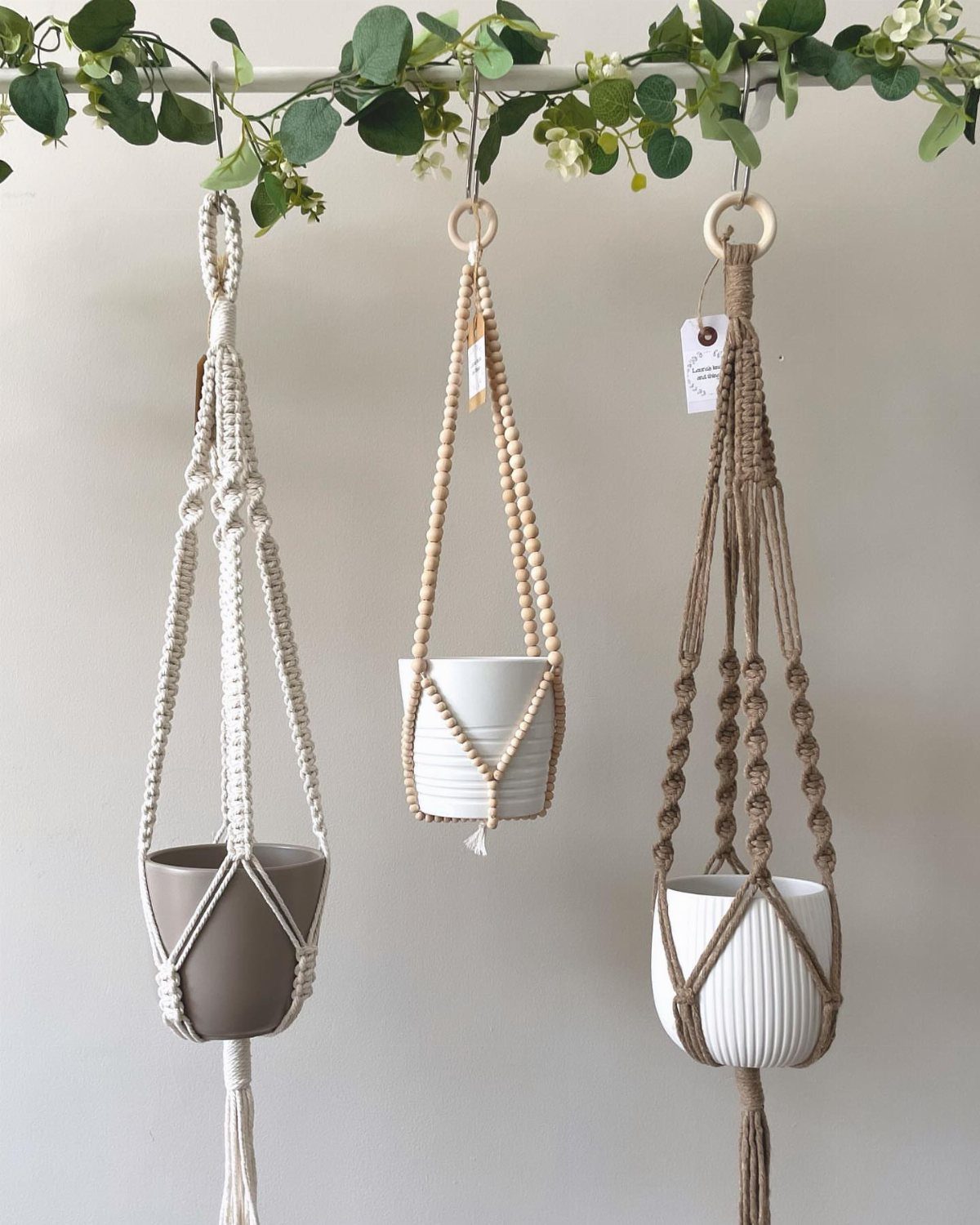 Three macramé plant hangers holding ceramic pots hanging from a leafy vine rod.