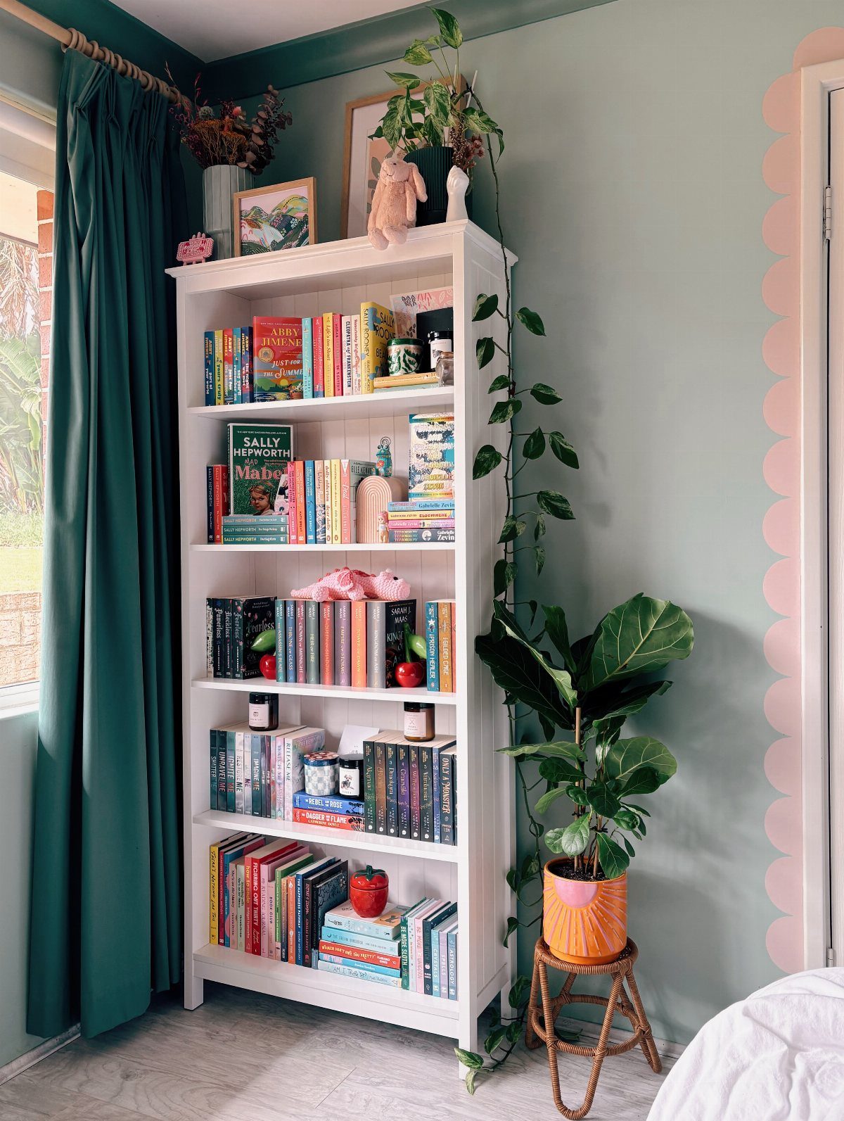 White bookshelf with colorful books stacked vertically and horizontally next to green plants.