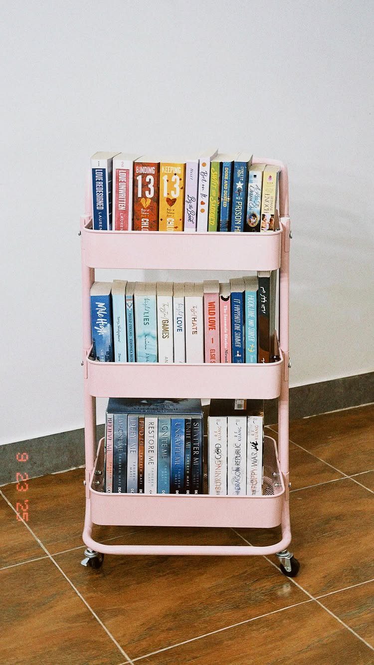 Pink three-tier rolling storage trolley filled with books against a white wall.
