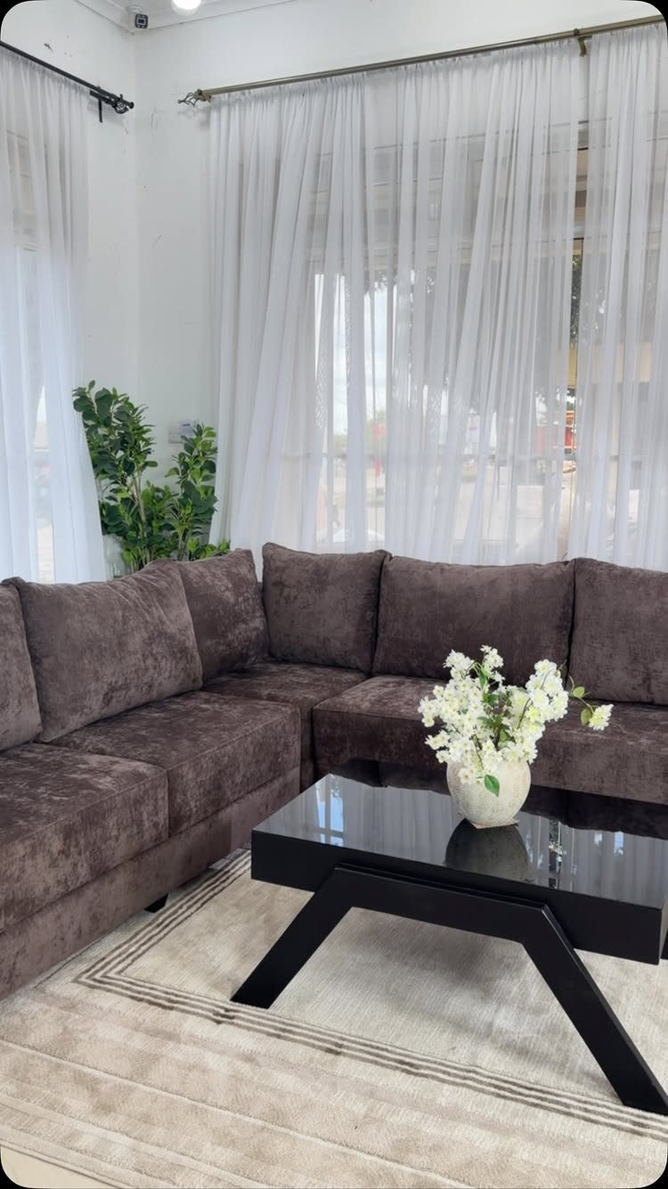 Dark brown sectional couch with a black glass coffee table and white curtains.