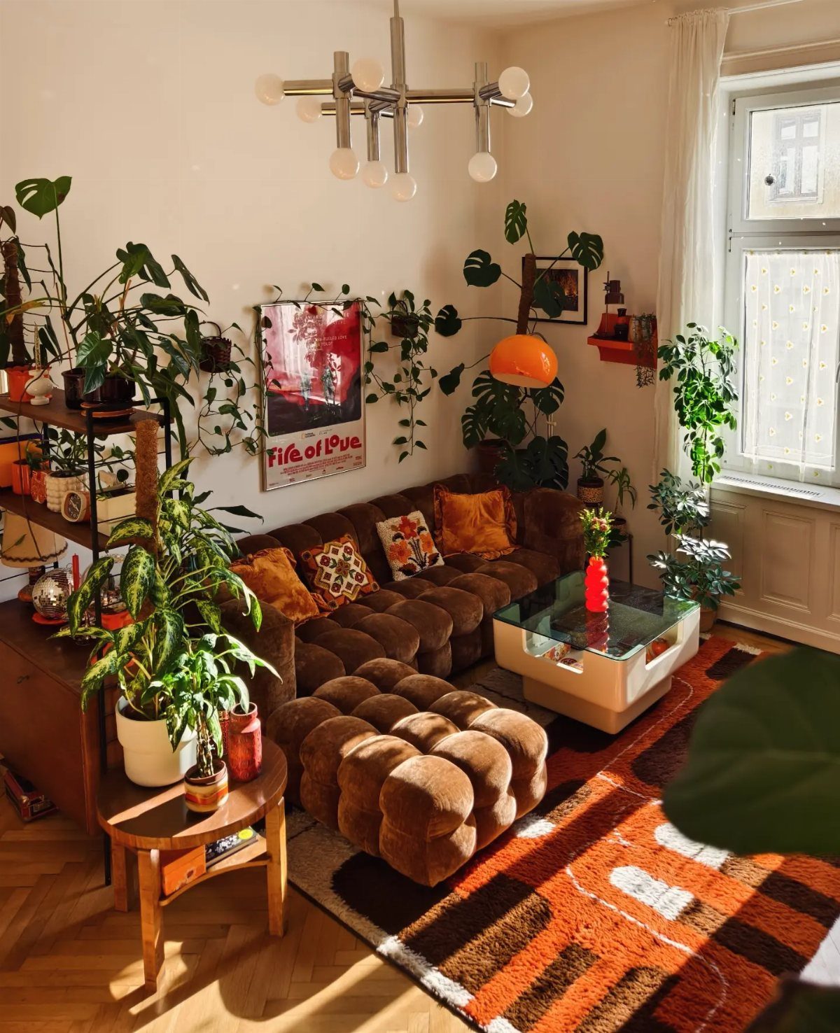 Brown velvet sofa and ottoman with a graphic orange rug and many indoor plants.