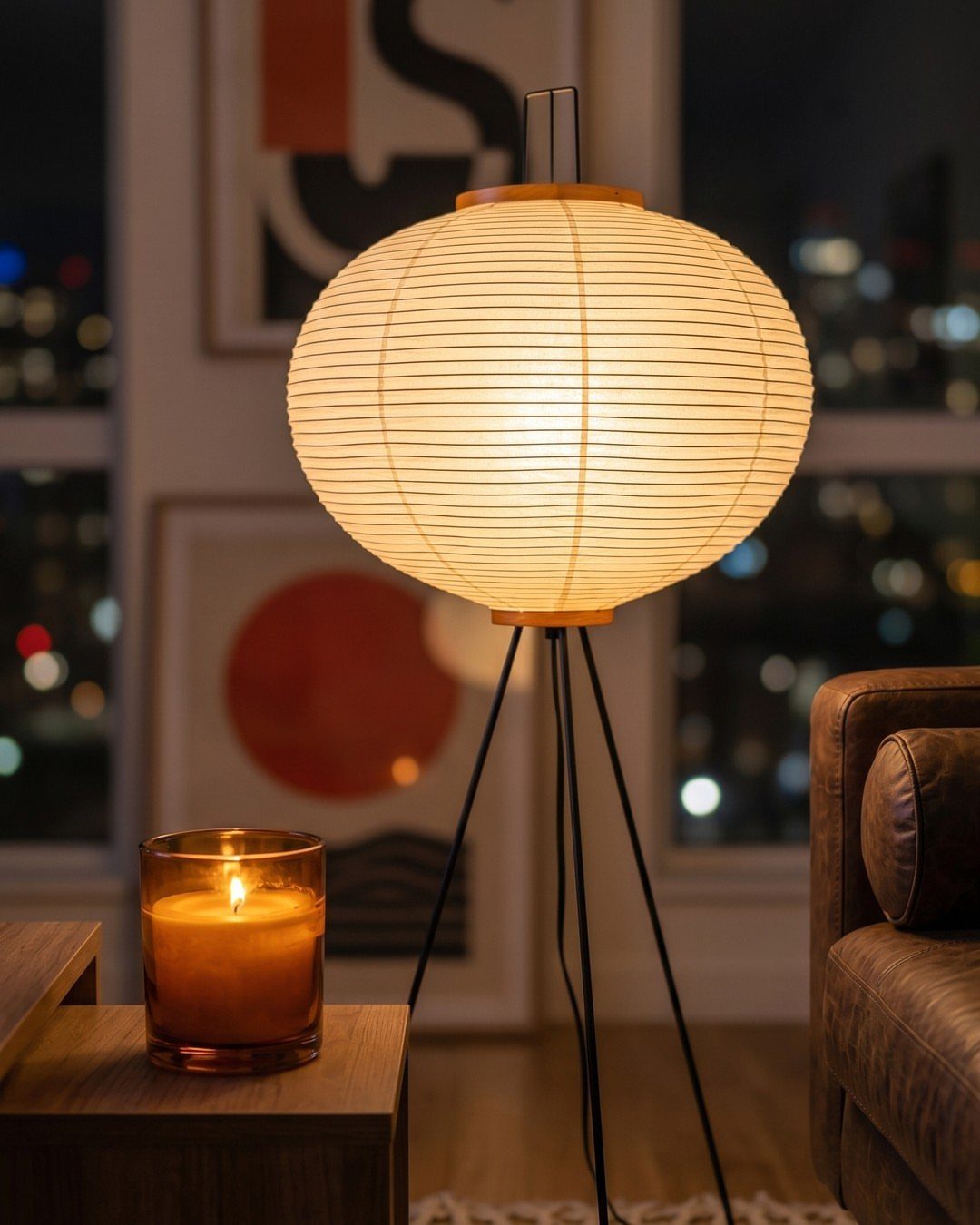 Glowing paper lantern floor lamp and lit candle beside a brown leather sofa.