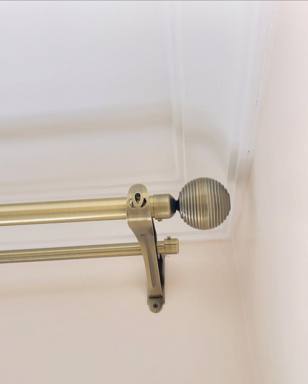 Brass double curtain rod with a ribbed spherical finial against a white wall.