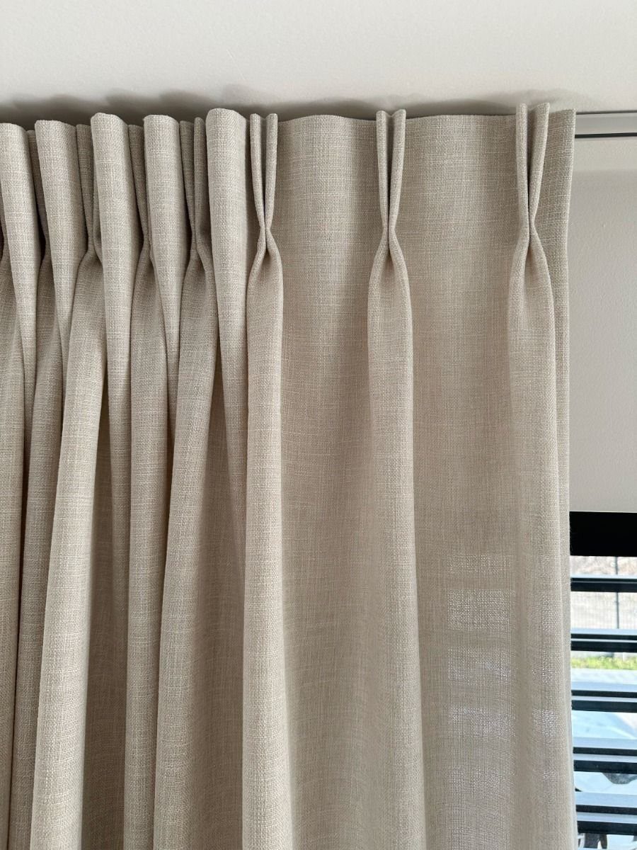 Beige linen pleated curtains hanging from a ceiling track in a bright room.