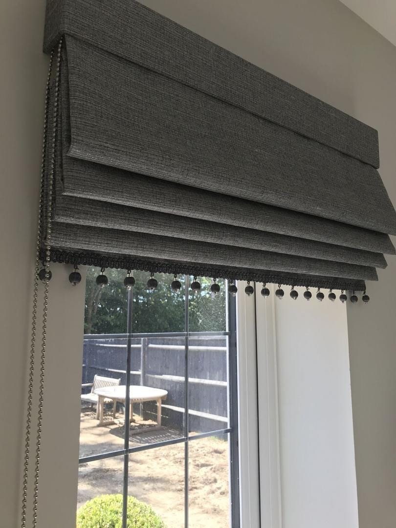 Grey fabric Roman blind with beaded trim hanging over a living room window.