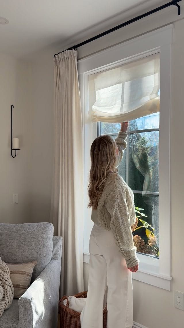 Woman adjusting cream roman shades and neutral curtains in a bright living room