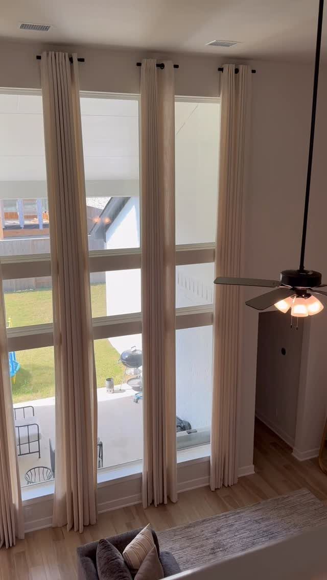 Cream-colored drapery panels hanging over large living room windows overlooking a green yard.