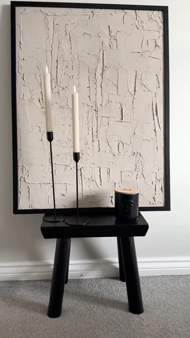 Textured white abstract painting leaning on a wall above a black bench with candles.