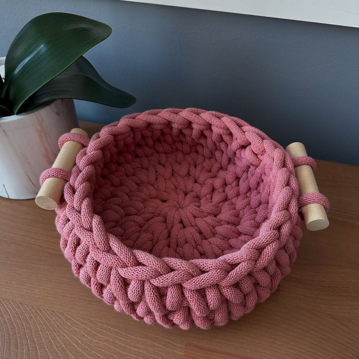 Pink chunky knit basket with wooden handles on a light wooden table.