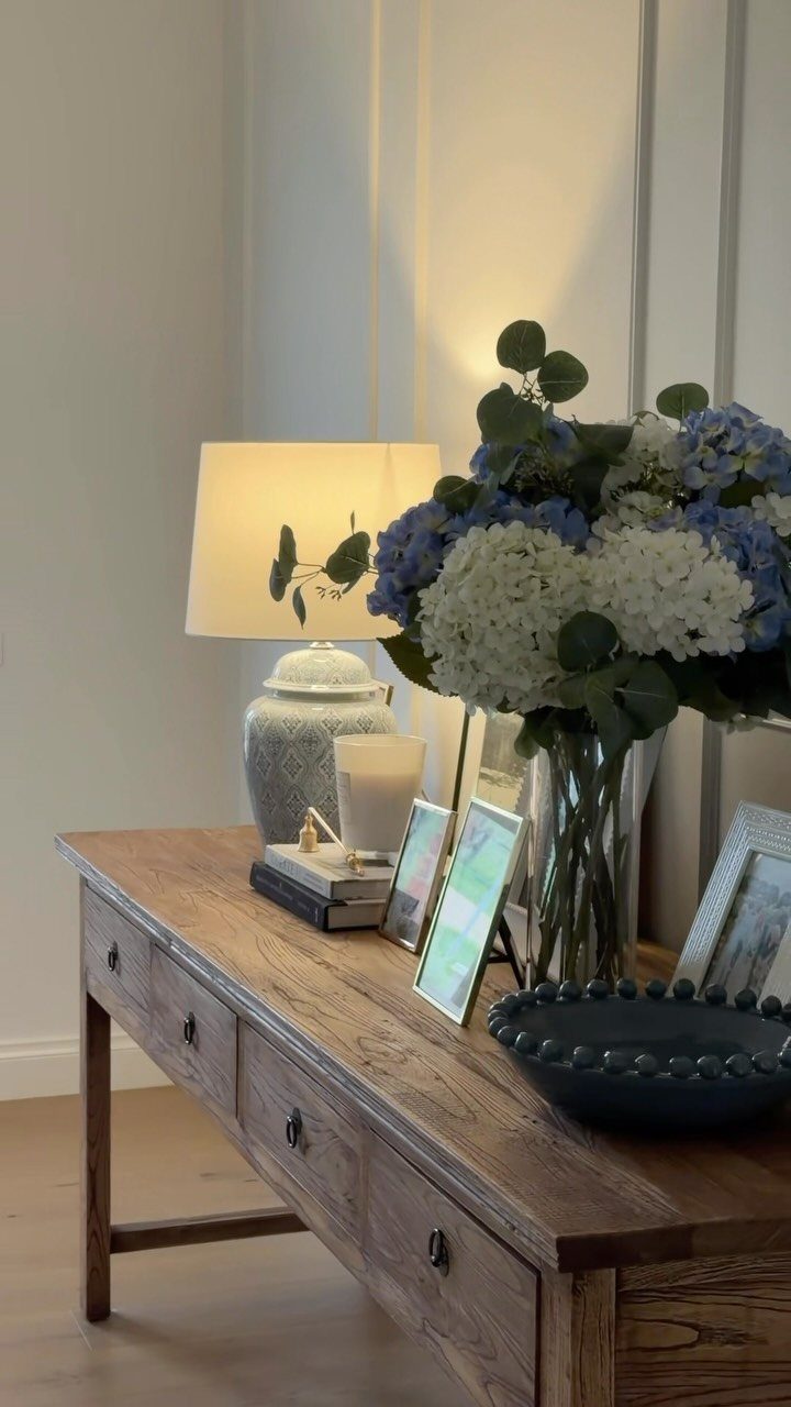 Rustic wooden console table with blue hydrangeas, a white lamp, and framed photos.