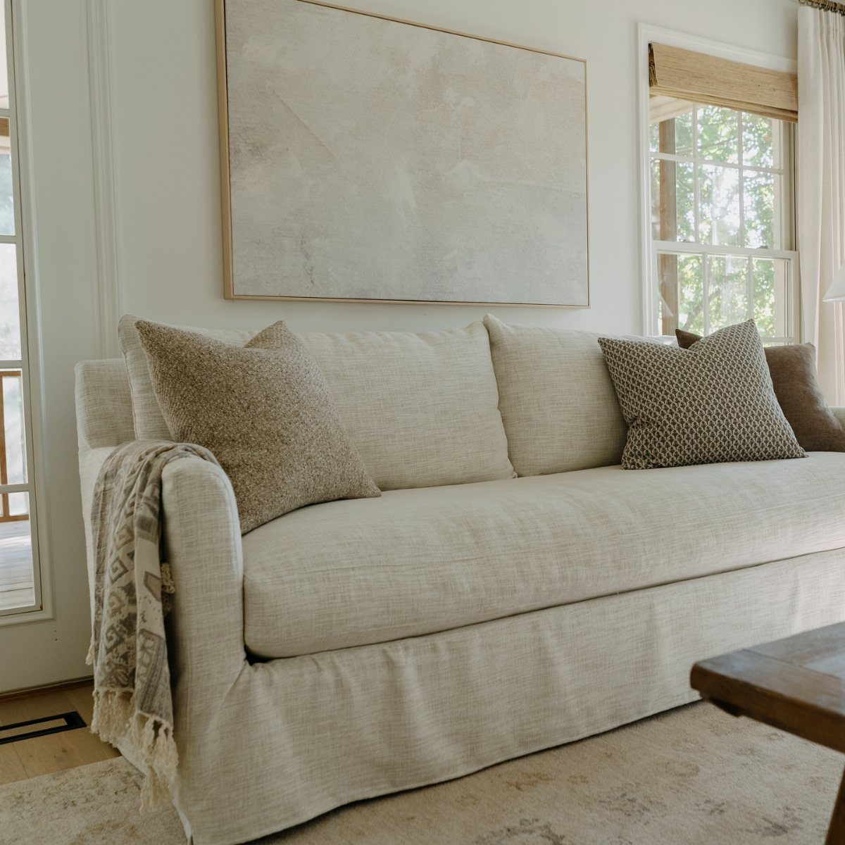 Cream slipcovered sofa with textured throw pillows and a blanket in a bright room.