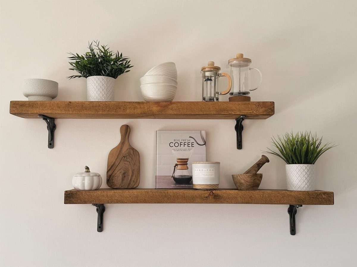 Two solid wood shelves with black brackets holding kitchenware and plants on white wall