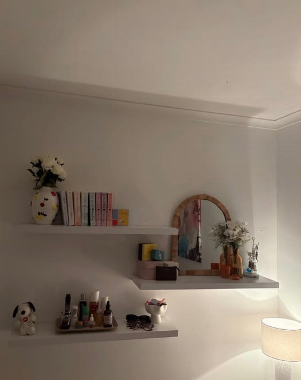 White floating shelves on a white wall holding books, flowers, and skincare products.