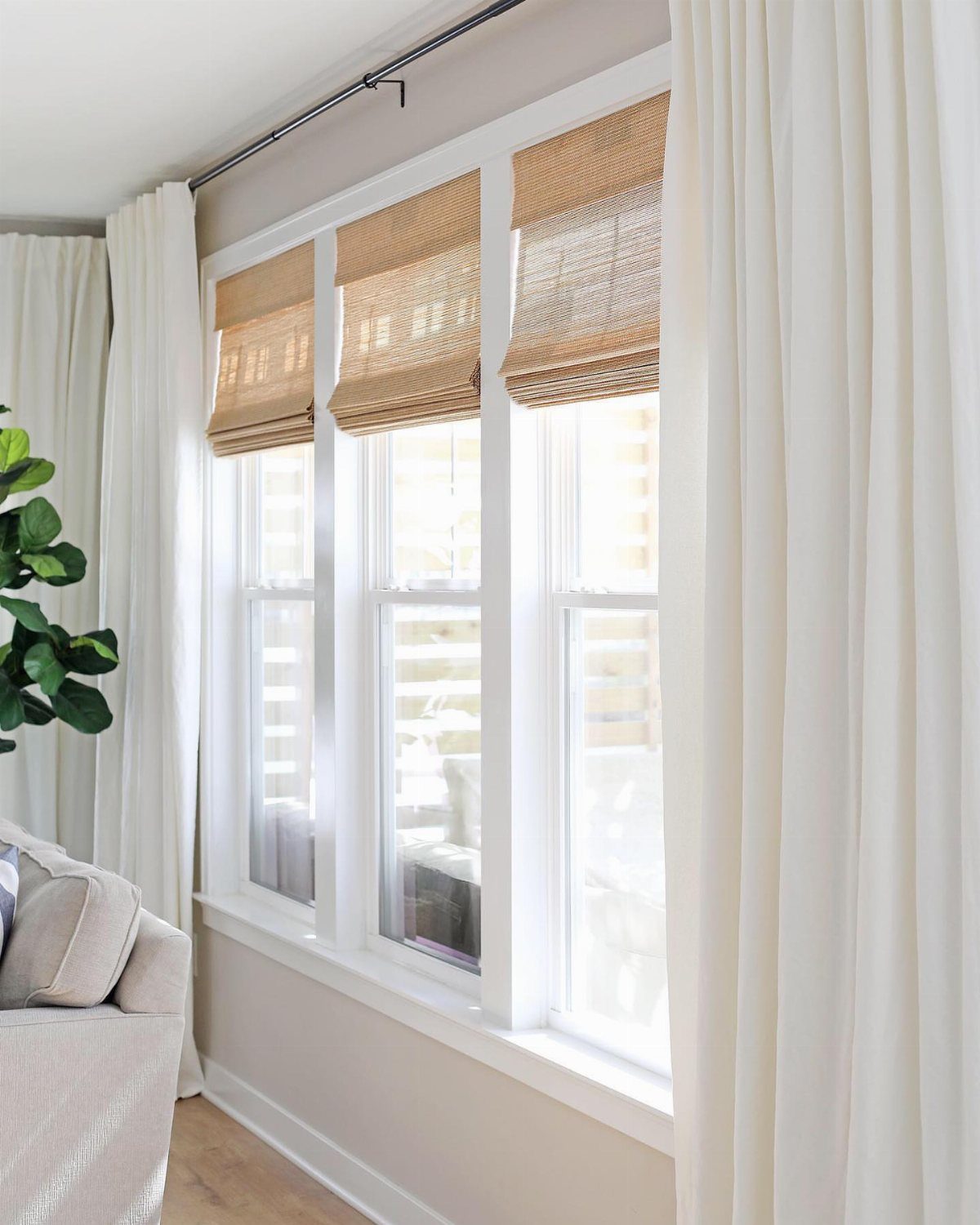Woven bamboo shades on white windows with white curtains and a beige sofa.