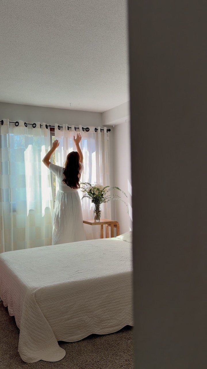 Woman adjusting white sheer curtains in a bright, minimalist white bedroom