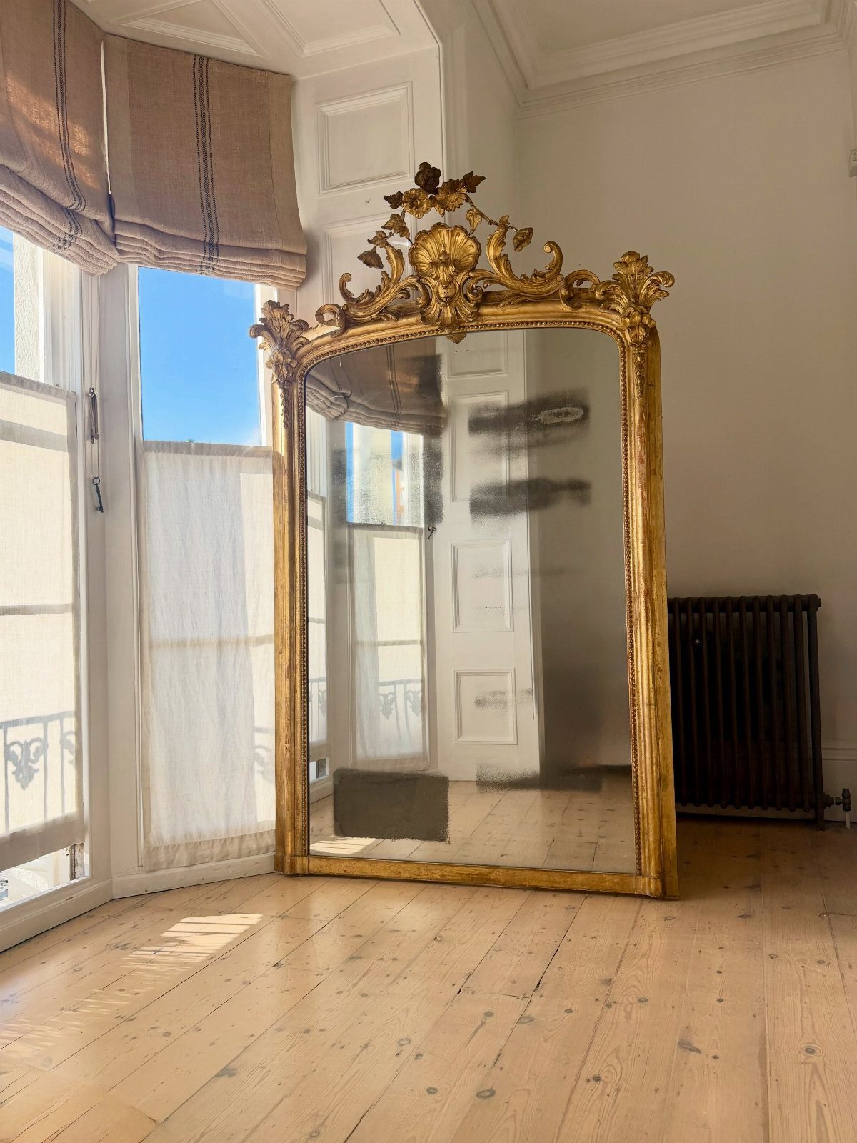 Ornate gold gilded mirror leaning against a white wall on light wood flooring.