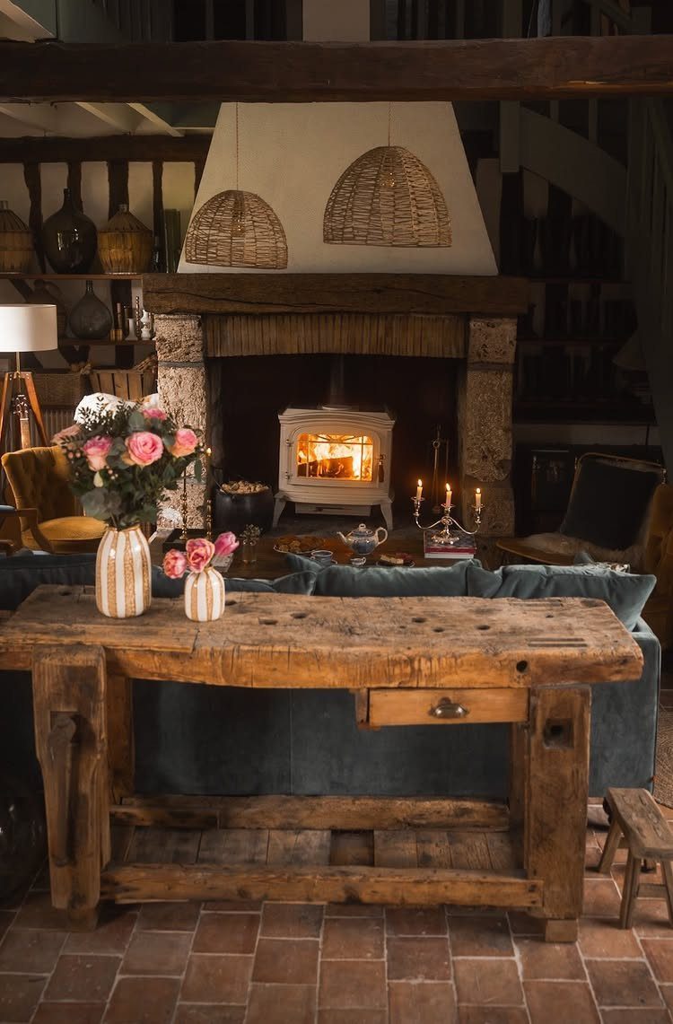 Rustic living room with wooden table, pink flowers, and glowing stone fireplace stove.