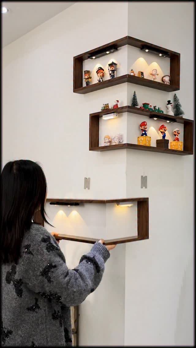 Woman installing brown wooden L-shaped corner shelves with built-in lights on white walls.