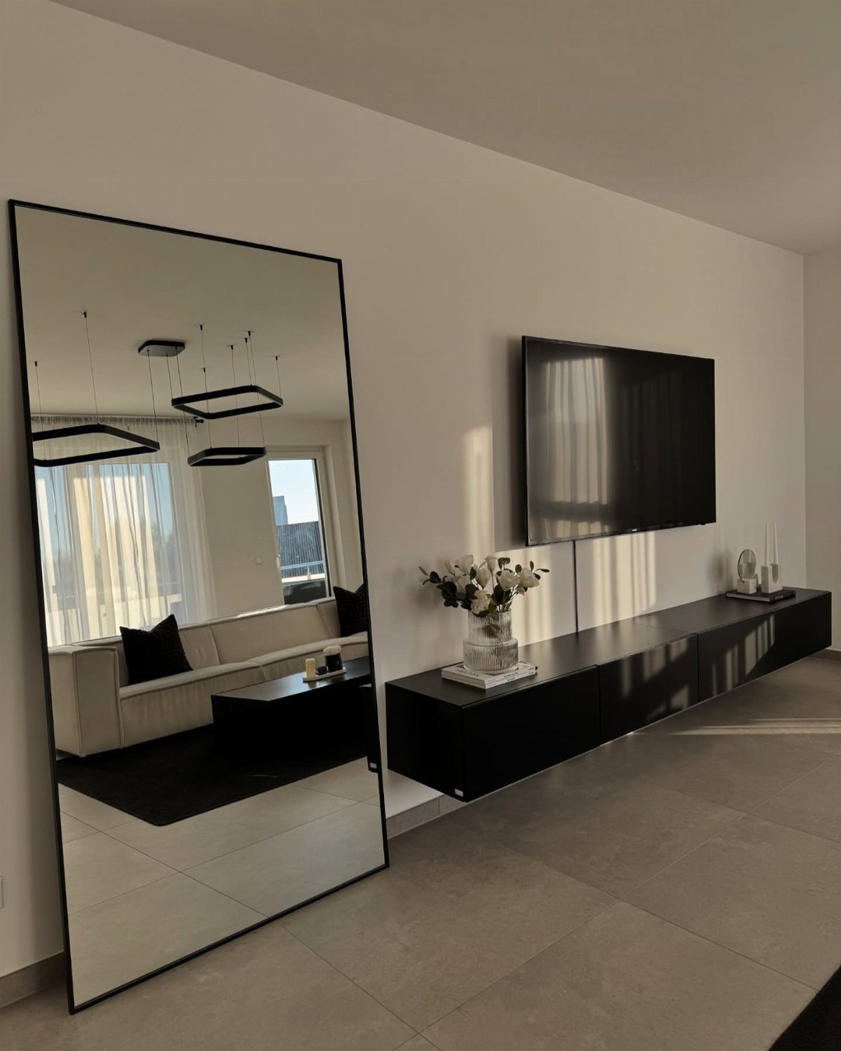 Large black-framed floor mirror reflecting a window and sofa in a modern room