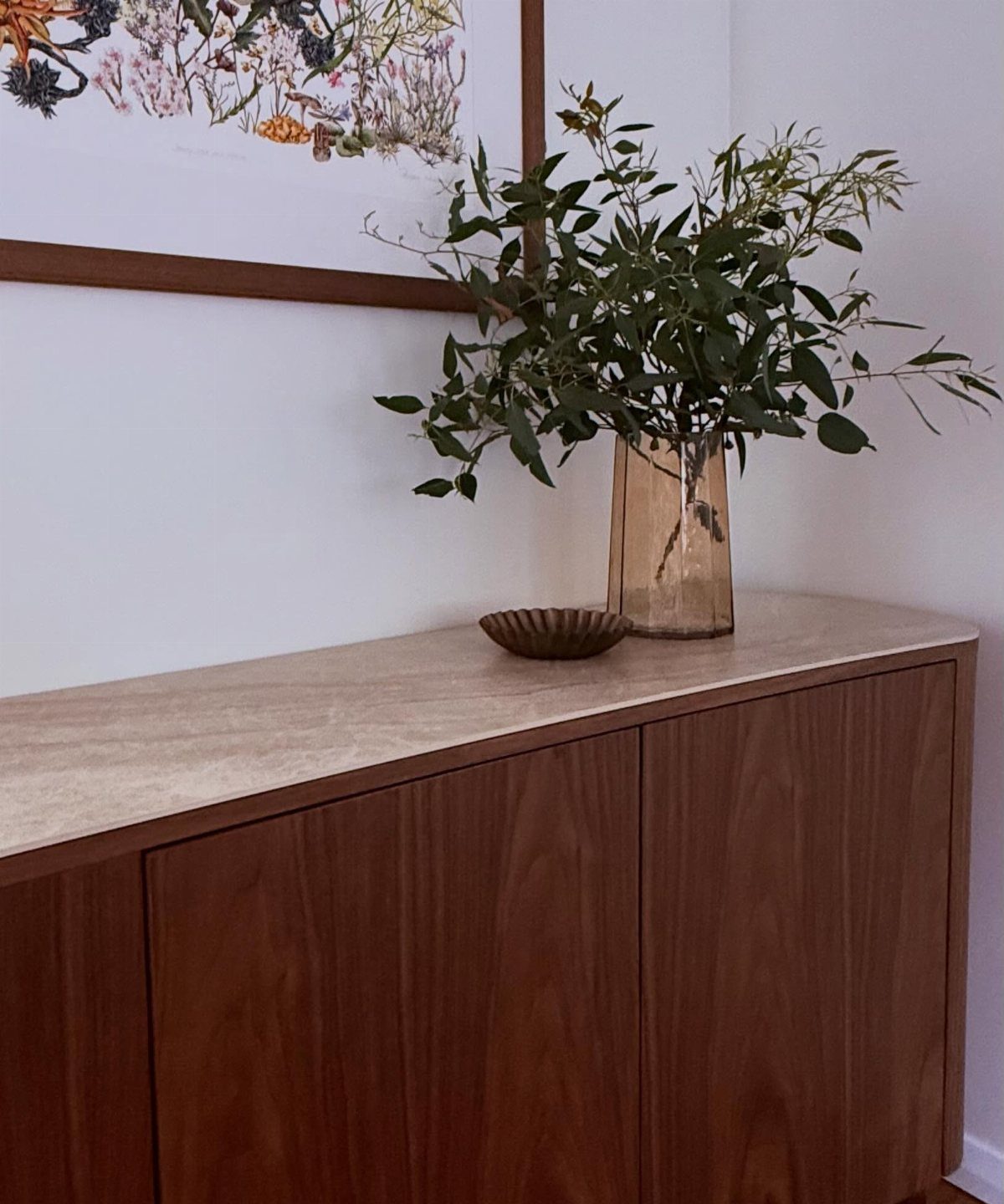 Wooden sideboard with stone top, glass vase with greenery, and framed wall art.