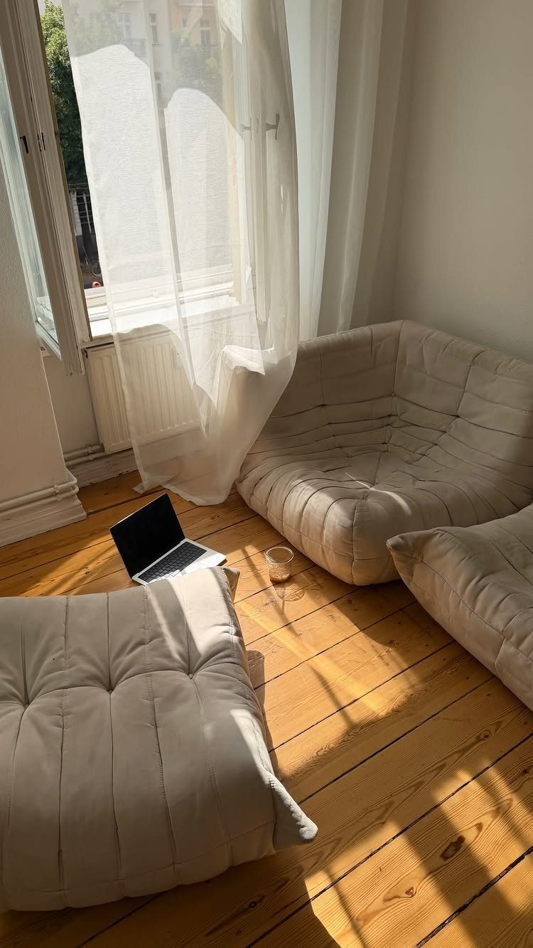 Beige tufted floor cushions and a laptop on light wood flooring by a window.