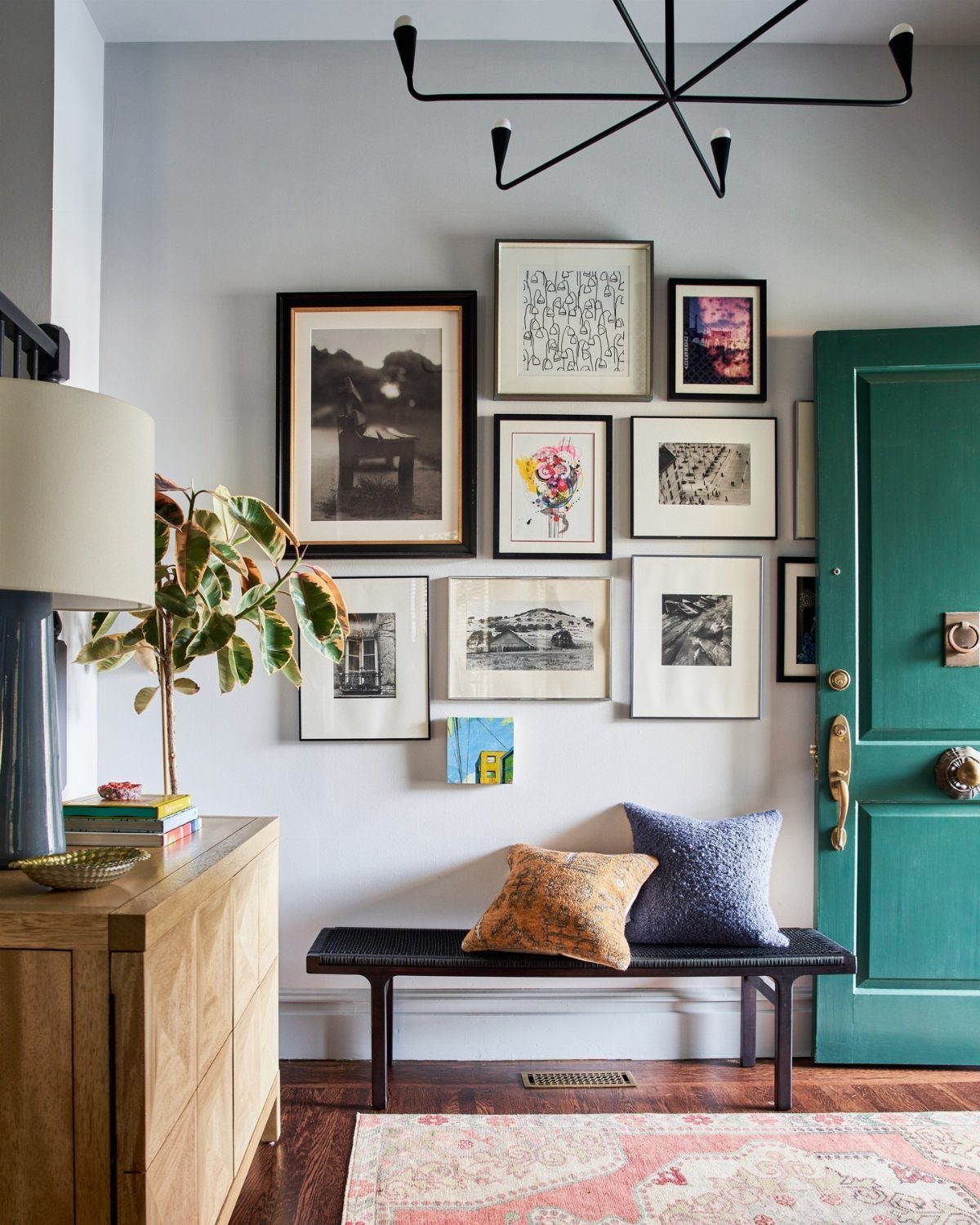 Asymmetrical gallery wall with mixed frames above a black bench and teal door.