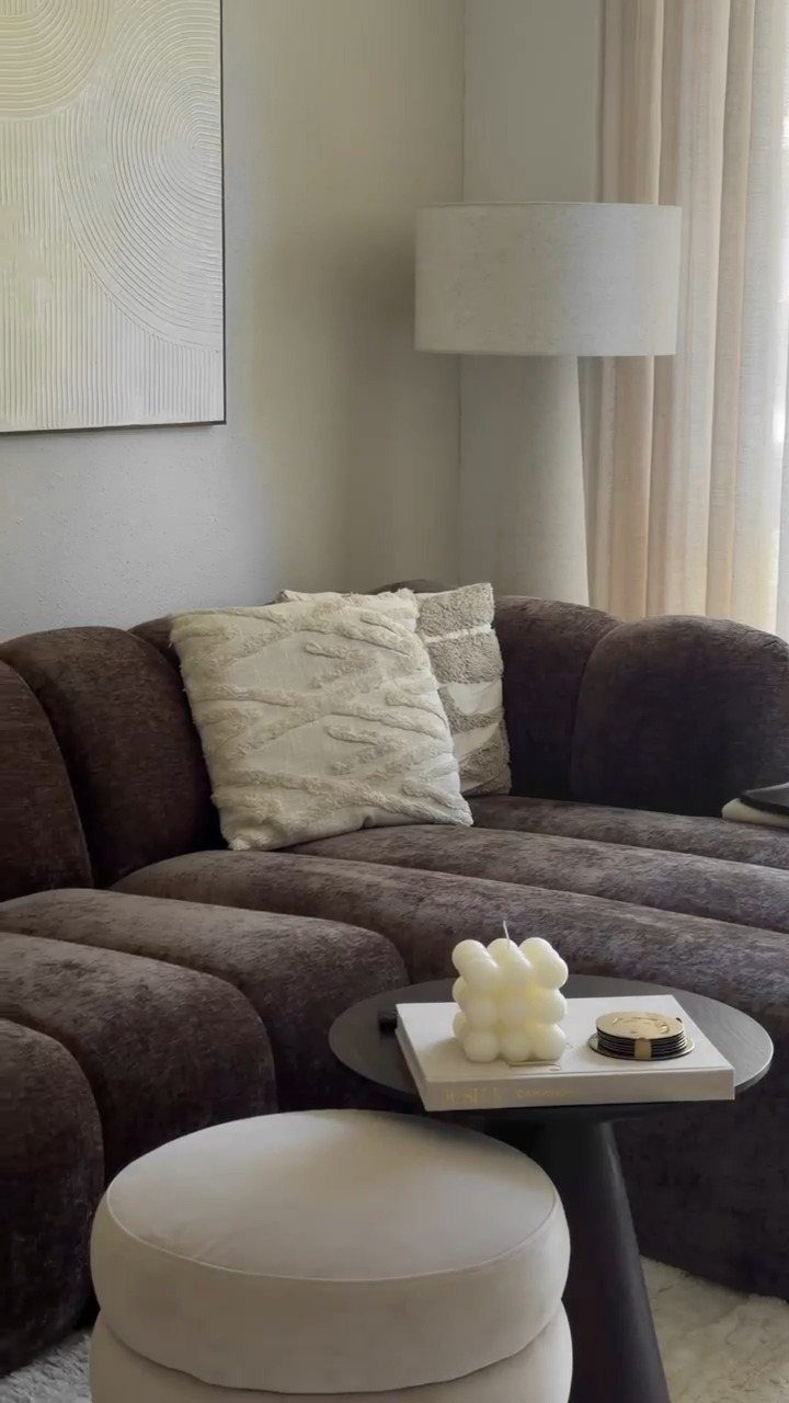 Brown curved sofa with cream pillows and a white ottoman in neutral living room