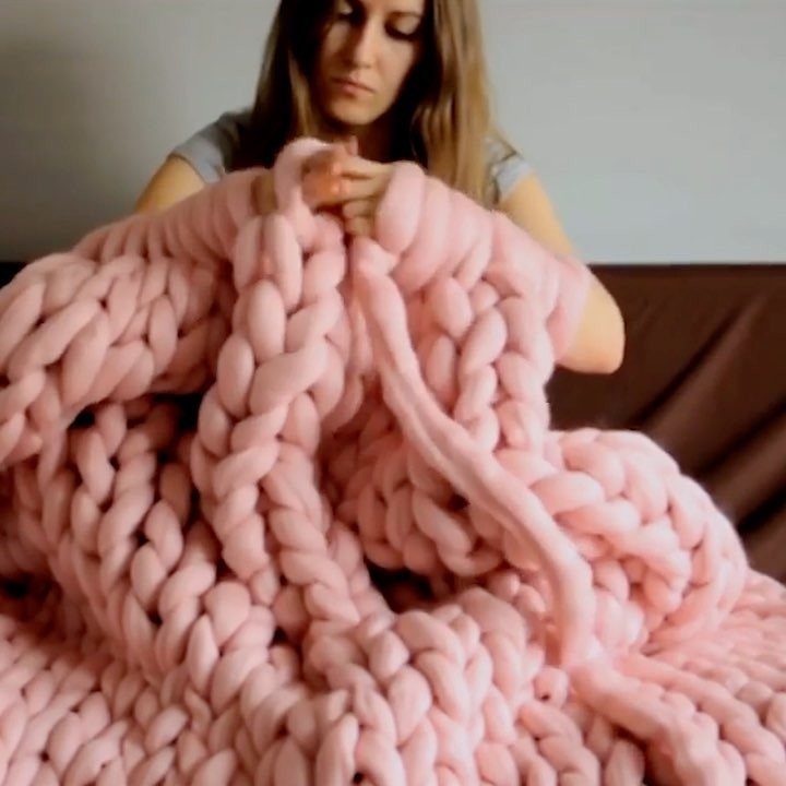 Woman holding a thick, chunky knit pink blanket over a brown sofa.