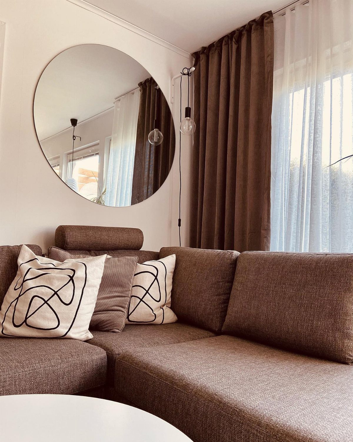Brown sectional sofa with patterned pillows, round wall mirror, and brown curtains.