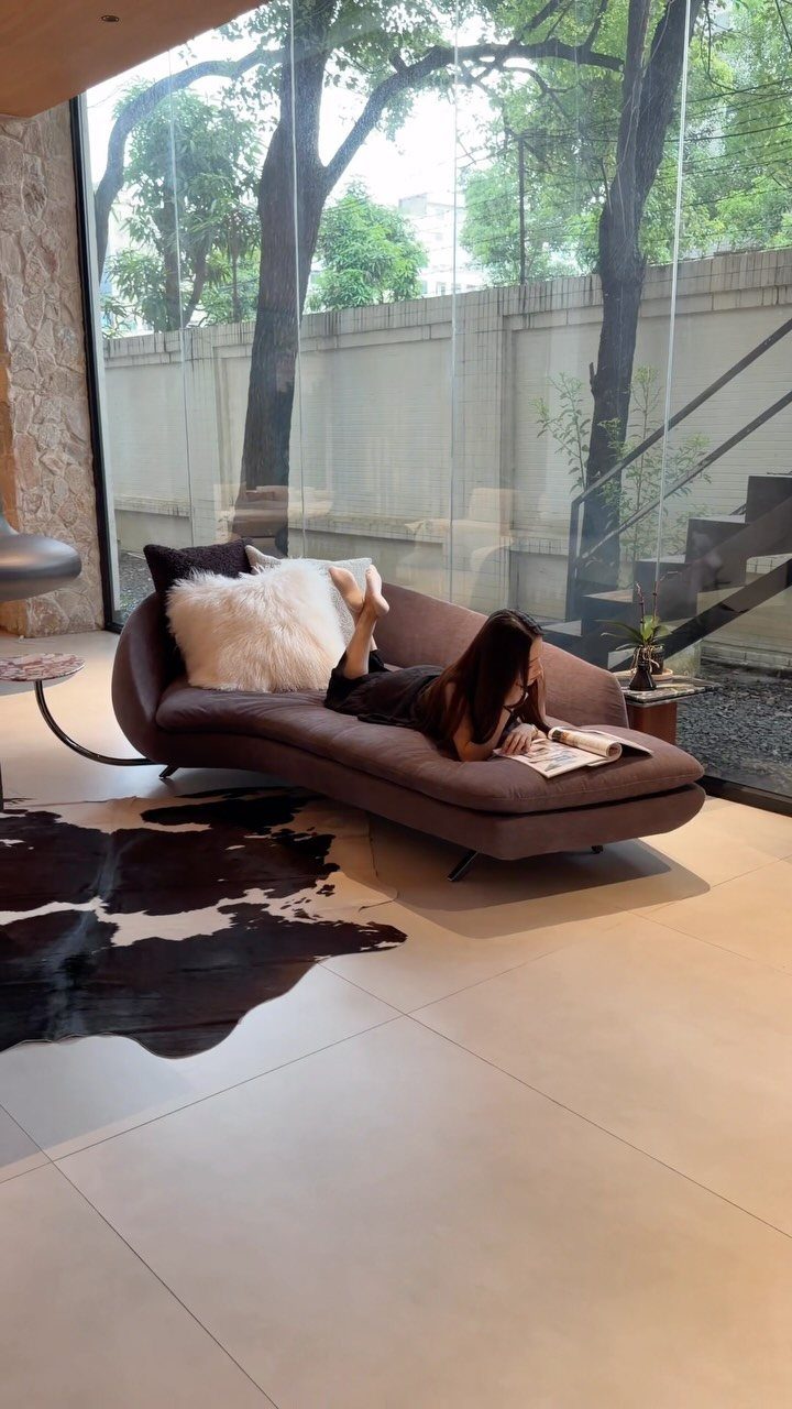 Woman relaxing on a brown curved chaise lounge in a modern living room.