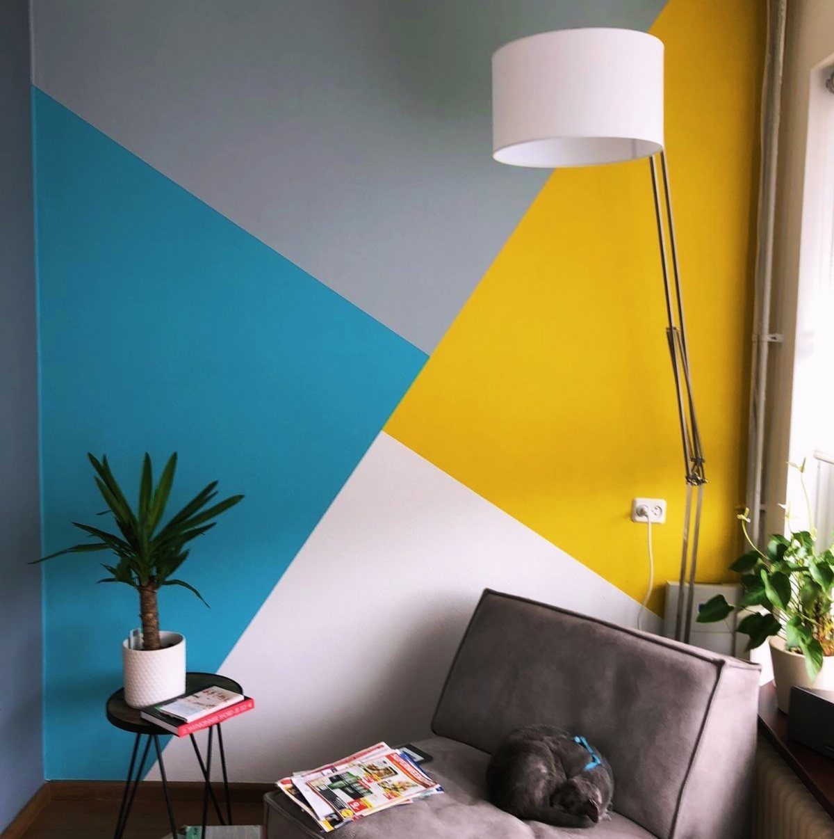 Geometric painted wall in blue, yellow, and grey behind a grey sofa and lamp.