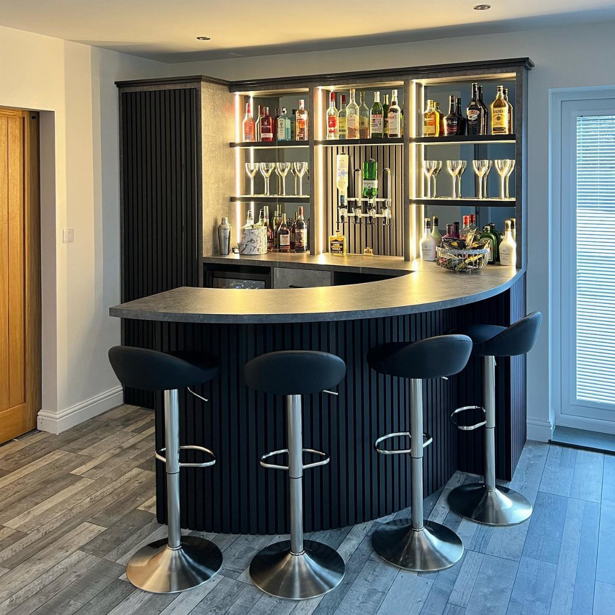 Modern home bar with curved dark counter, illuminated liquor shelves, and black bar stools.