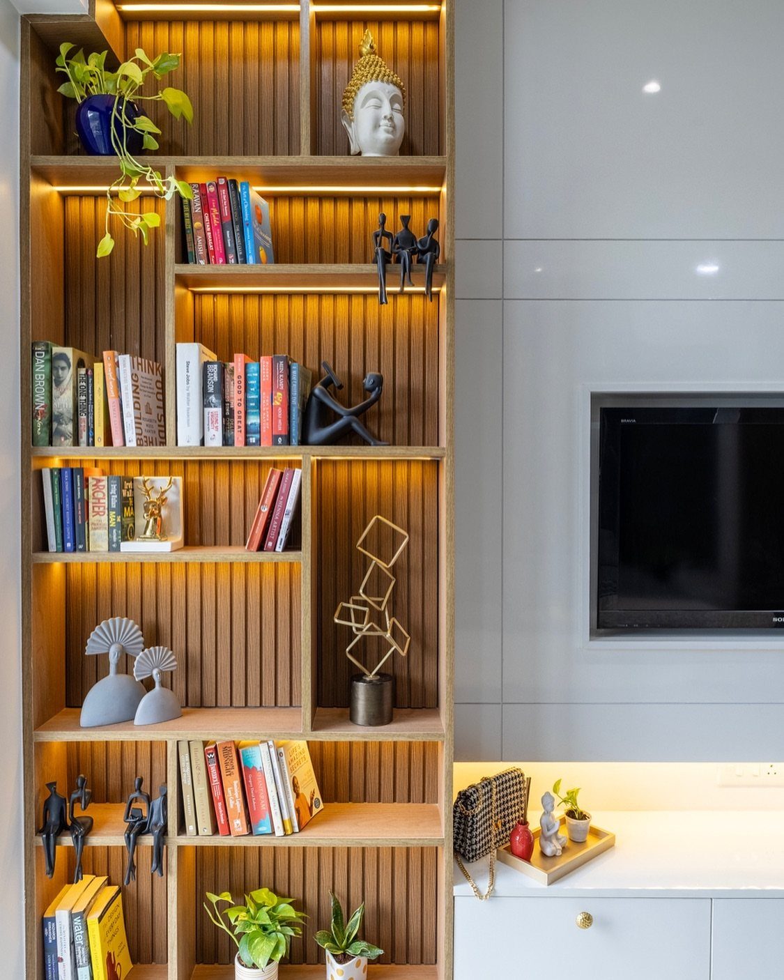 Tall wooden bookshelf with integrated lighting containing books, plants, and decorative sculptures.