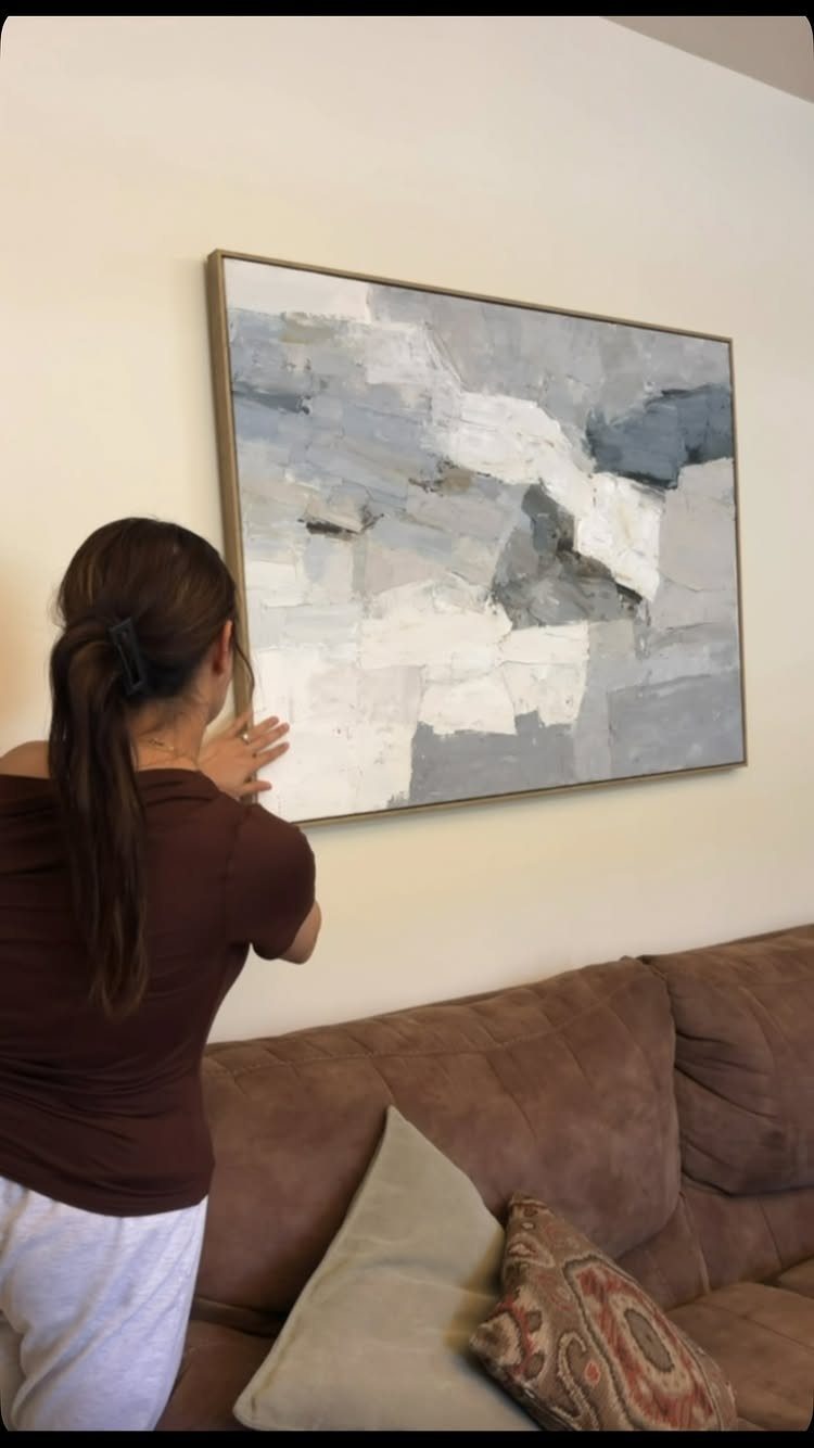 Woman hanging a large abstract painting with grey and white tones on a wall