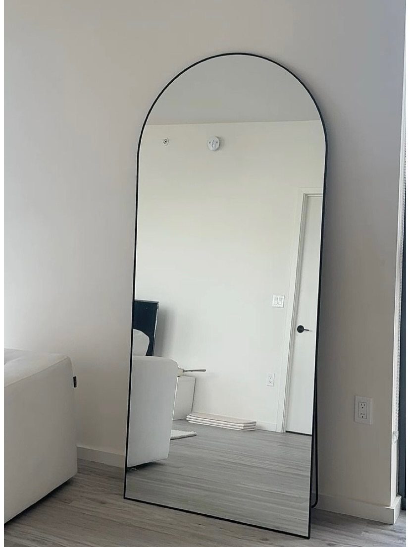 Arched full-length mirror with black frame leaning against a white living room wall.