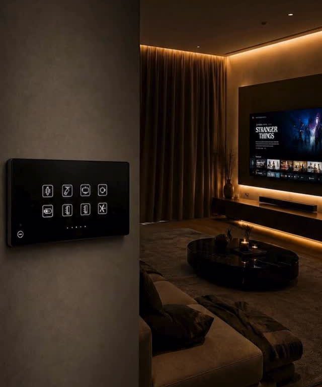 Digital control panel on a wall overlooking a living room with warm cove lighting.