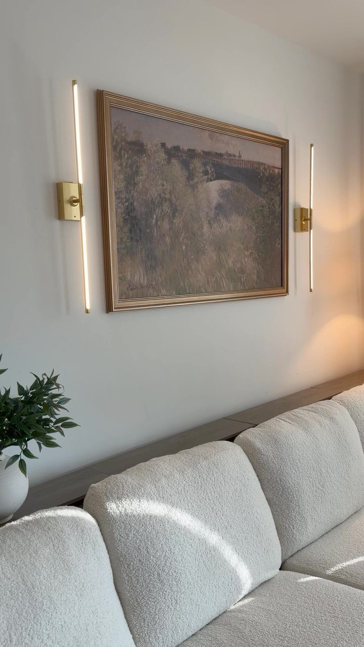 Two vertical gold wall sconces flanking a framed landscape painting above a white sofa.