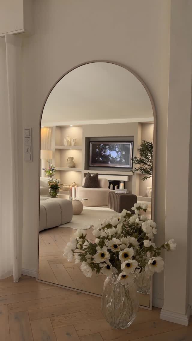 Arched floor mirror reflecting a neutral modern living room with white flowers foreground.