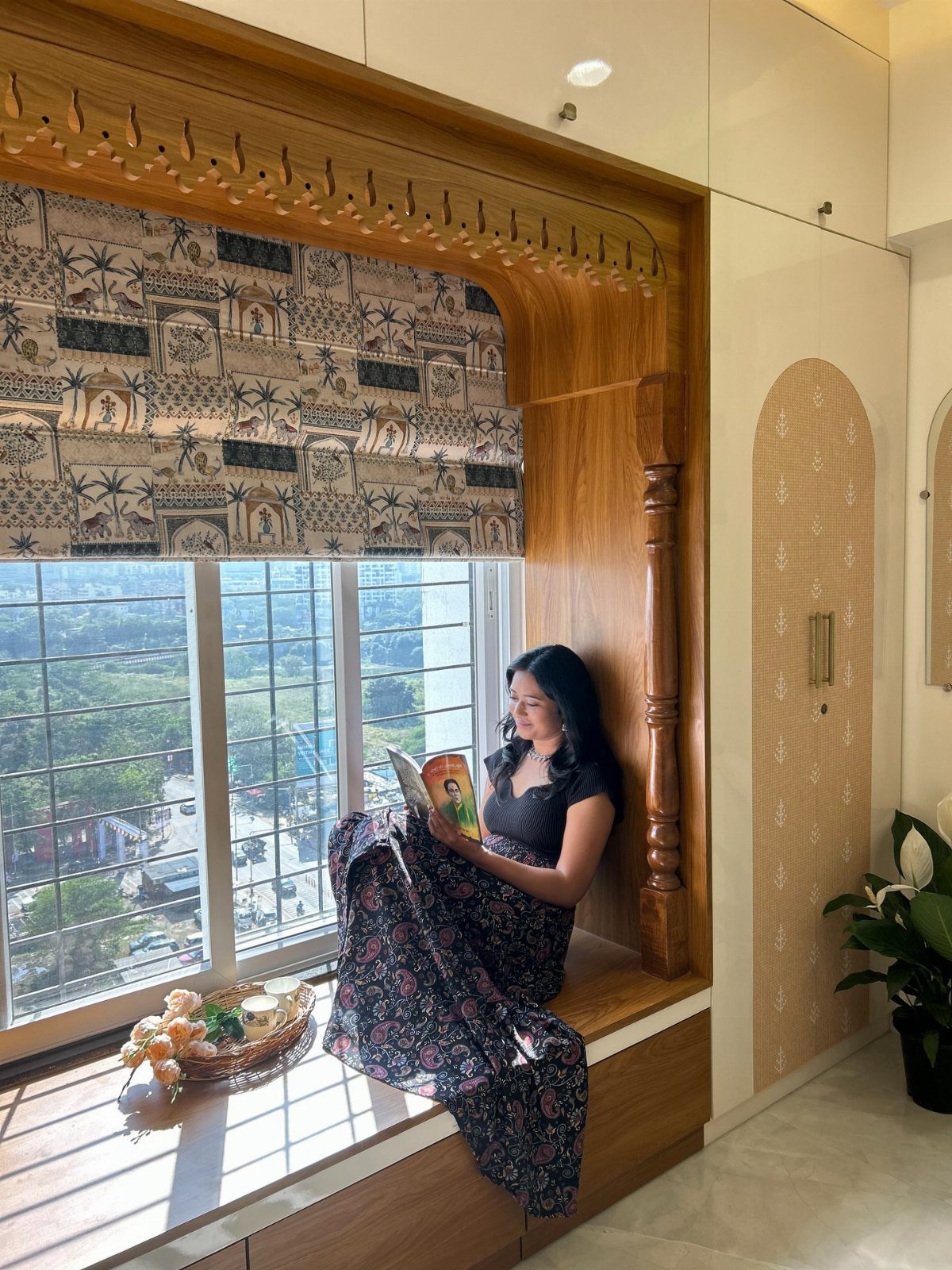 Woman reading a book on a carved wooden window seat with patterned blinds.