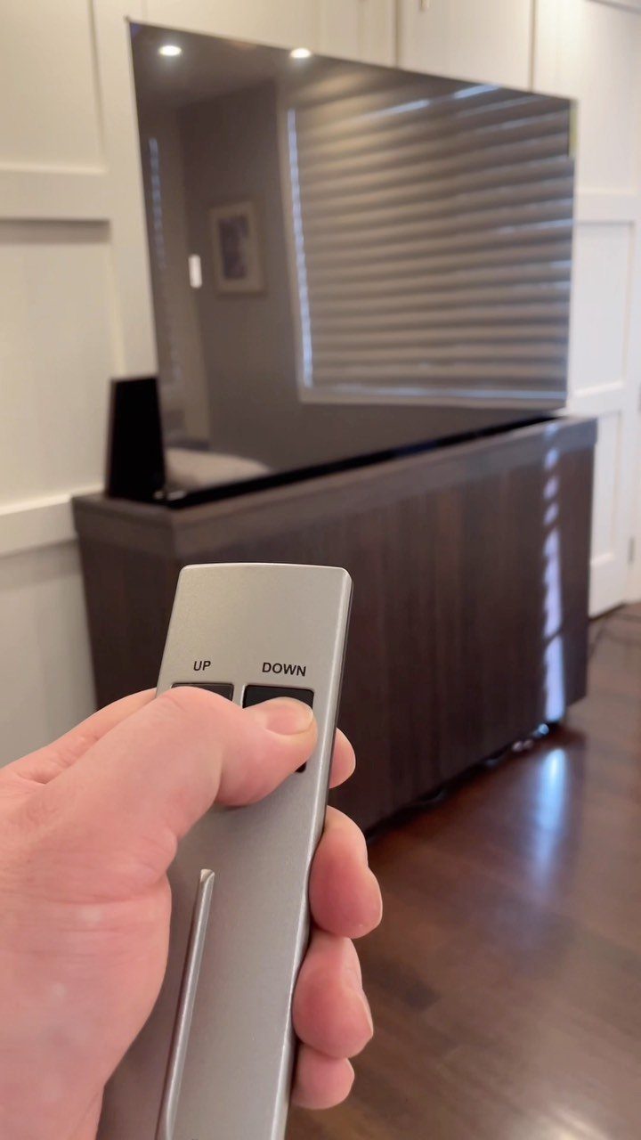 Hand holding a remote controlling a television on a dark wooden lift cabinet.
