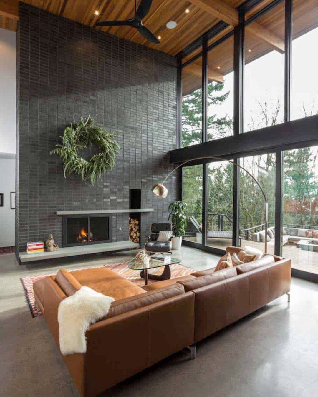 Living room with brown leather sofa and large floor-to-ceiling windows facing trees.