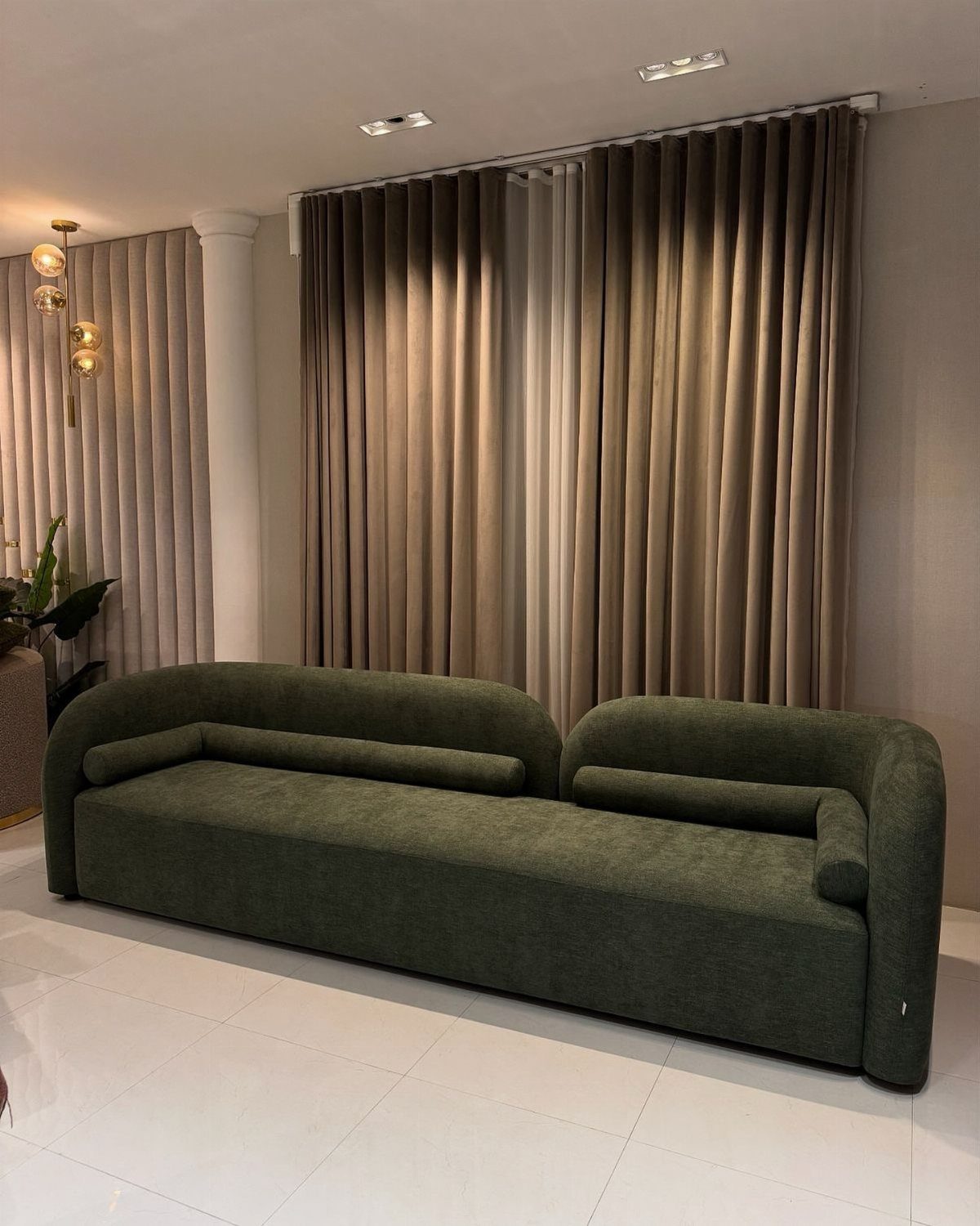 Dark green curved sofa in front of floor-to-ceiling beige pleated curtains