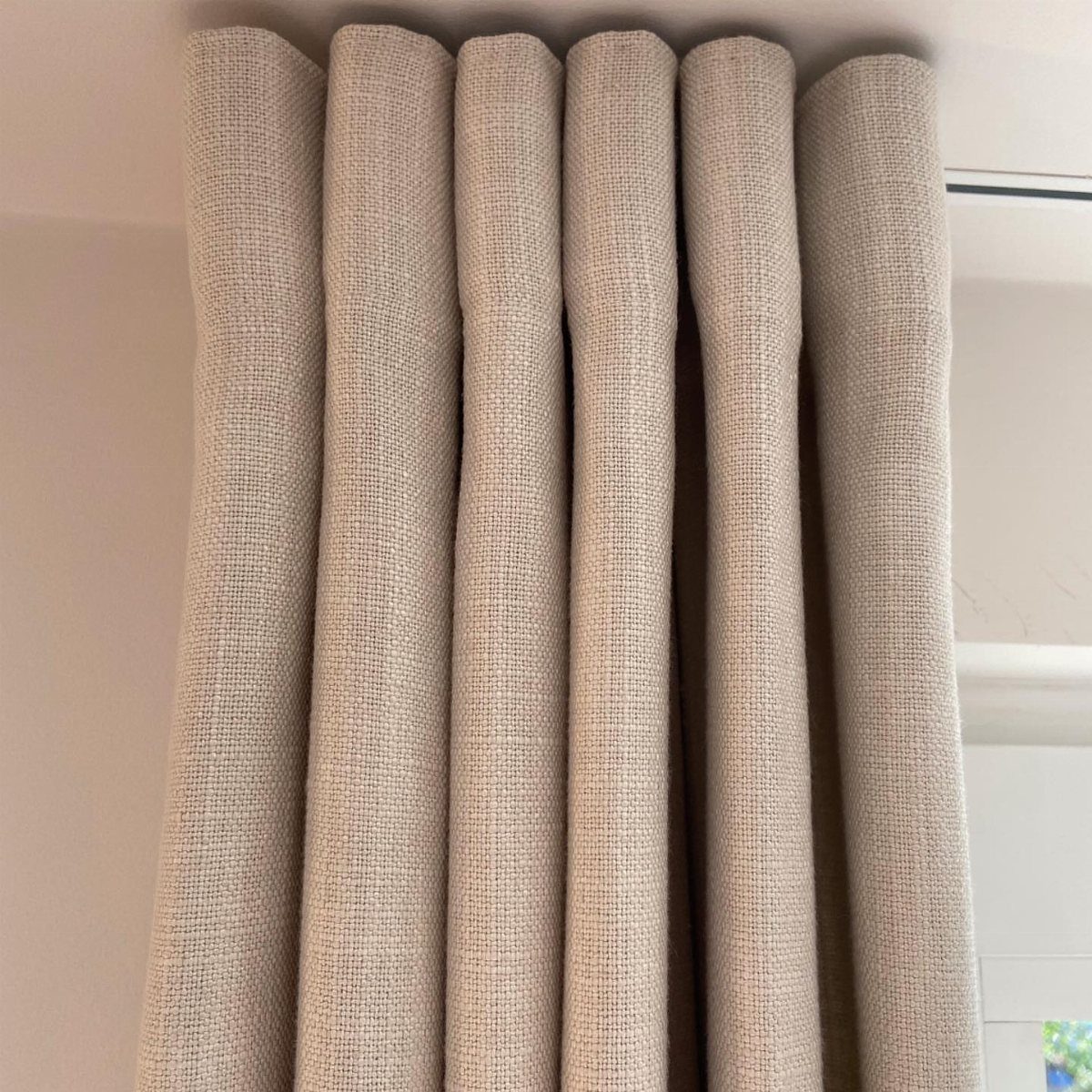 Beige textured fabric curtains hanging in deep vertical folds against a white wall.