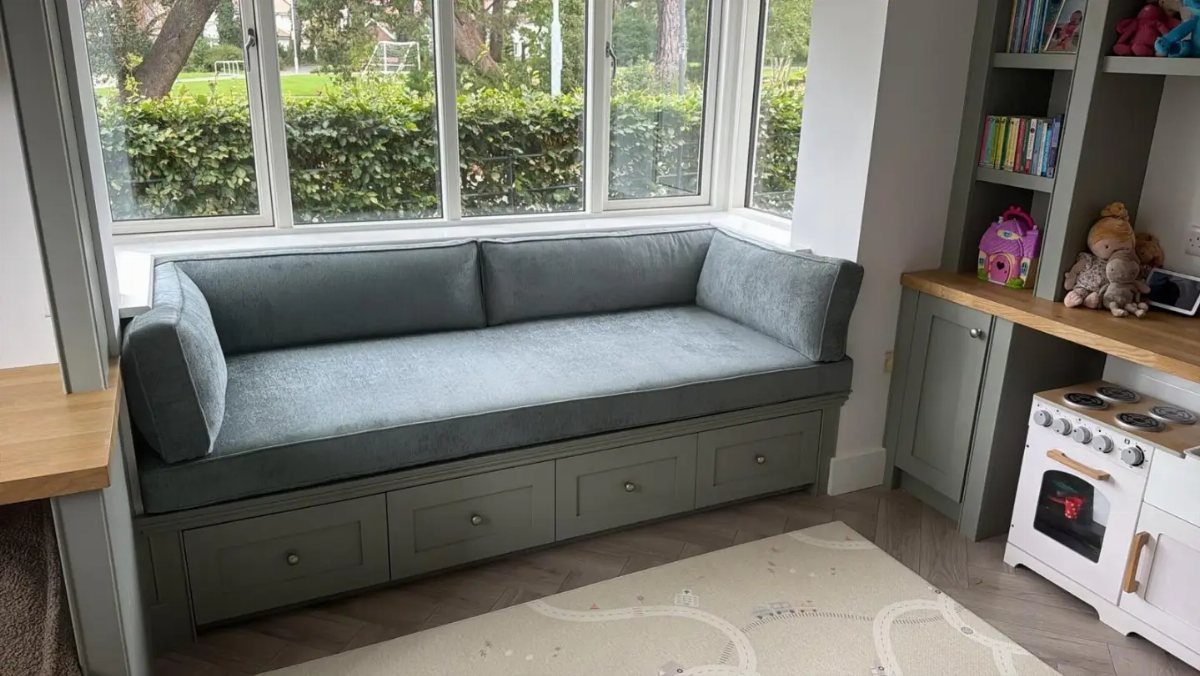 Grey built-in window seat couch with storage drawers beneath a large window.