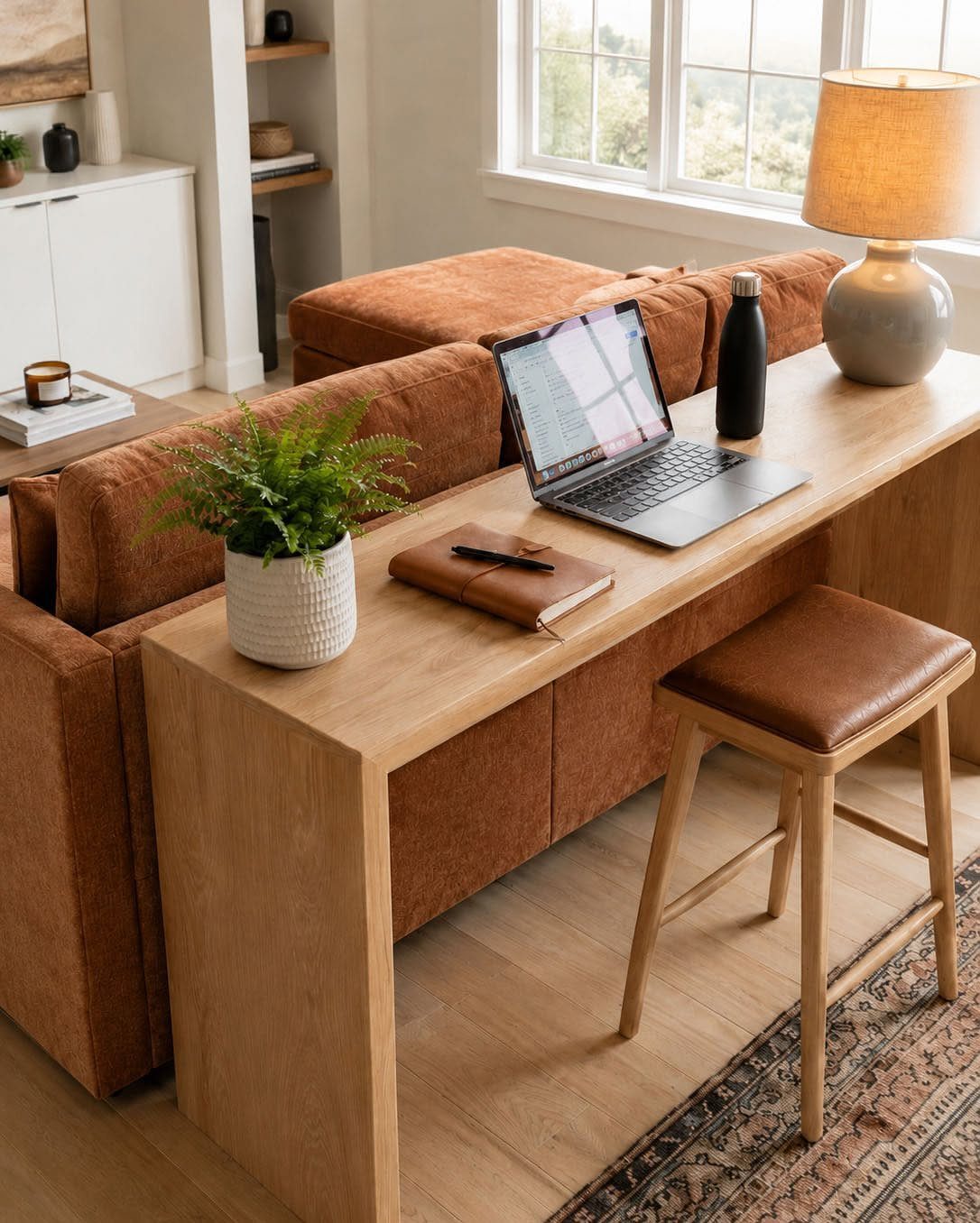 Light wood desk and brown leather stool positioned behind a brown fabric couch.