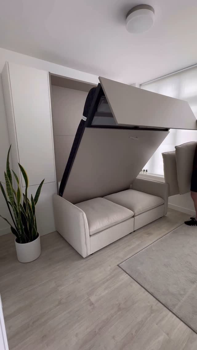 Beige sofa bed with a wall-mounted bed frame unfolding in a bright room.